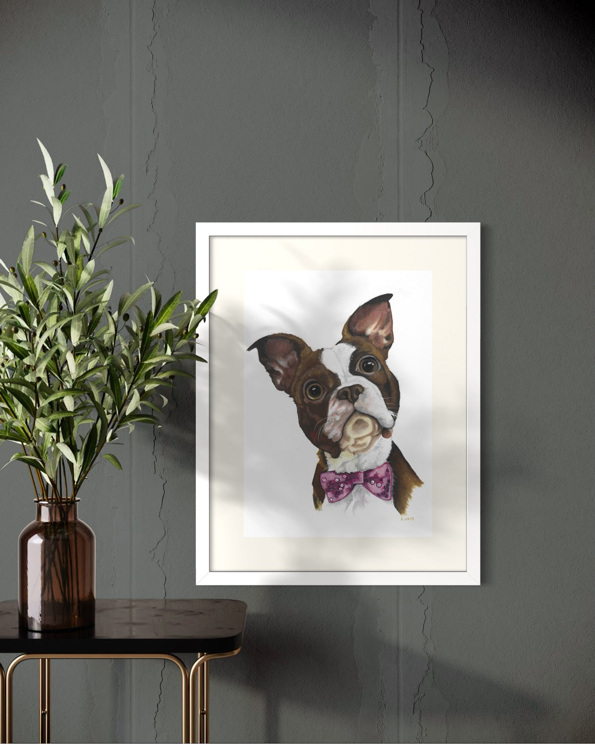 White framed A2 pet portrait commission on a dark charcoal wall beside a black brass-legged side table with olive branches in an amber glass vase.