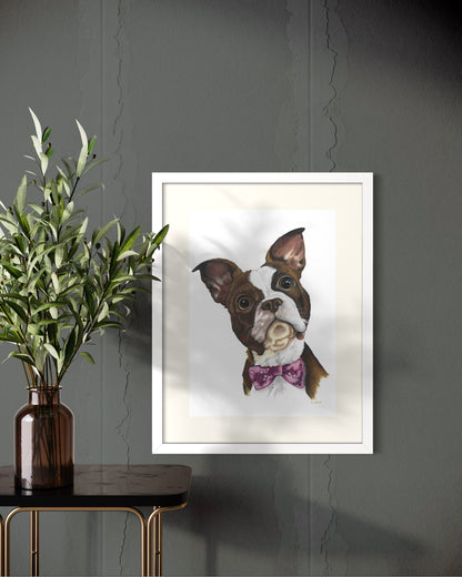 White framed A2 pet portrait commission on a dark charcoal wall beside a black brass-legged side table with olive branches in an amber glass vase.