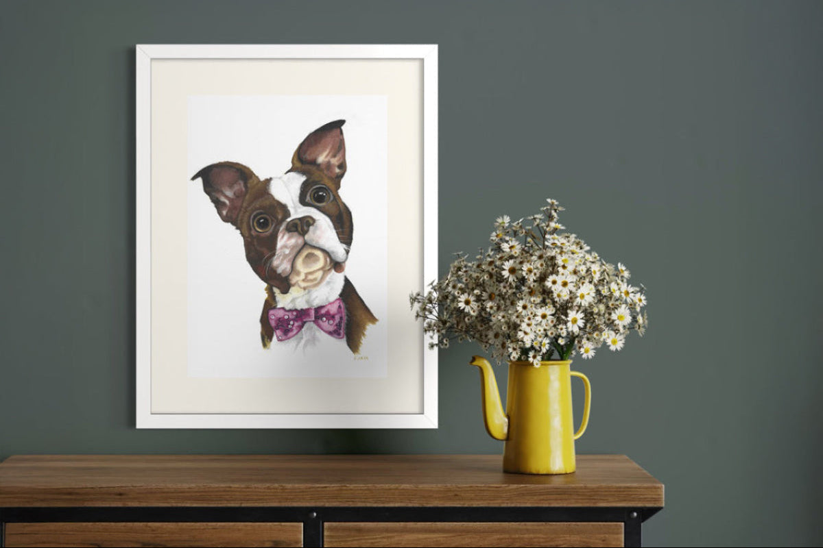 White framed A2 pastel pet portrait commission on a dark teal wall, beside an oak sideboard with a yellow enamel watering can and white daisies.
