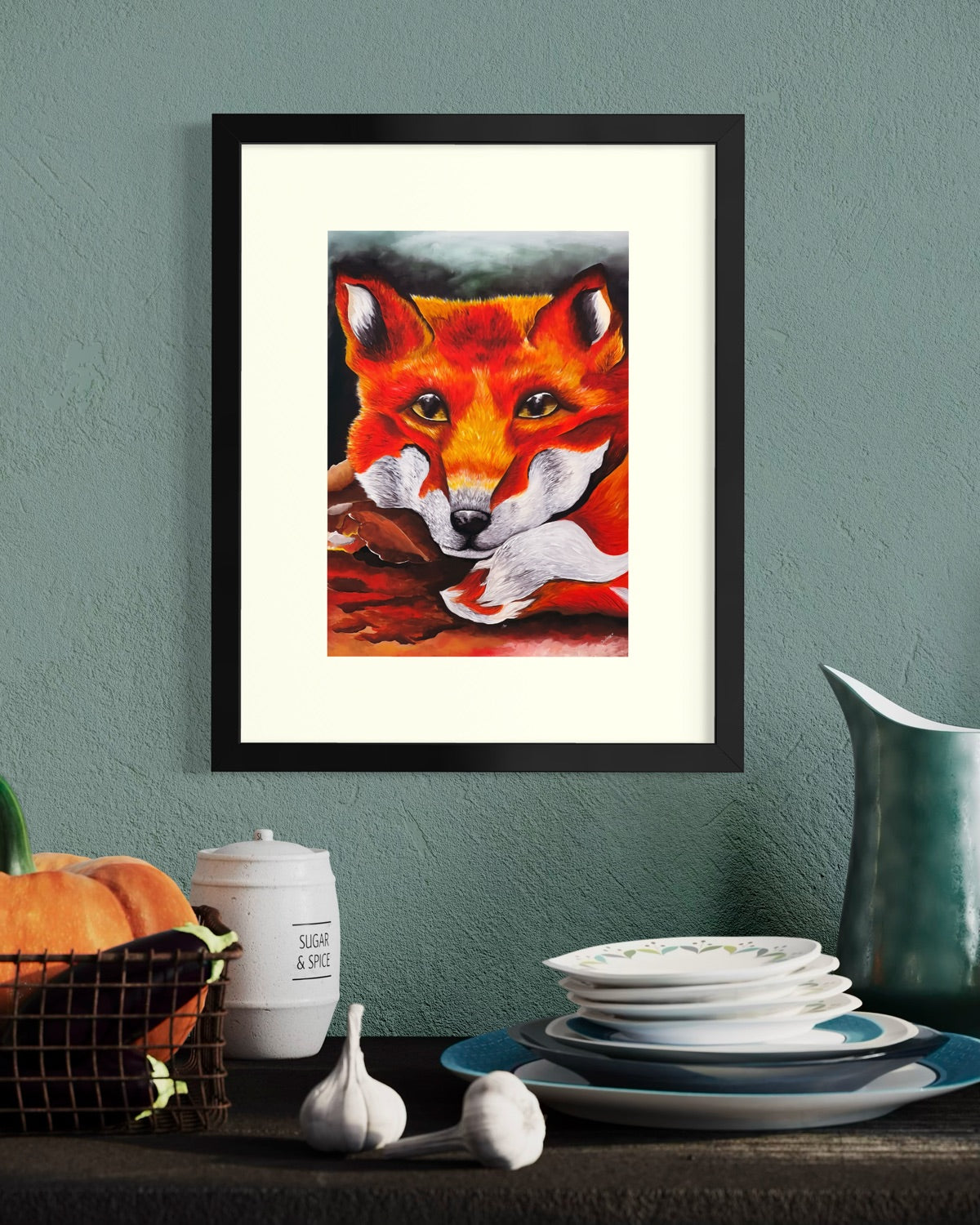 Black framed A4 red fox painting on a dusty teal kitchen wall above a wire basket of pumpkin and aubergine, sugar jar and stack of plates.