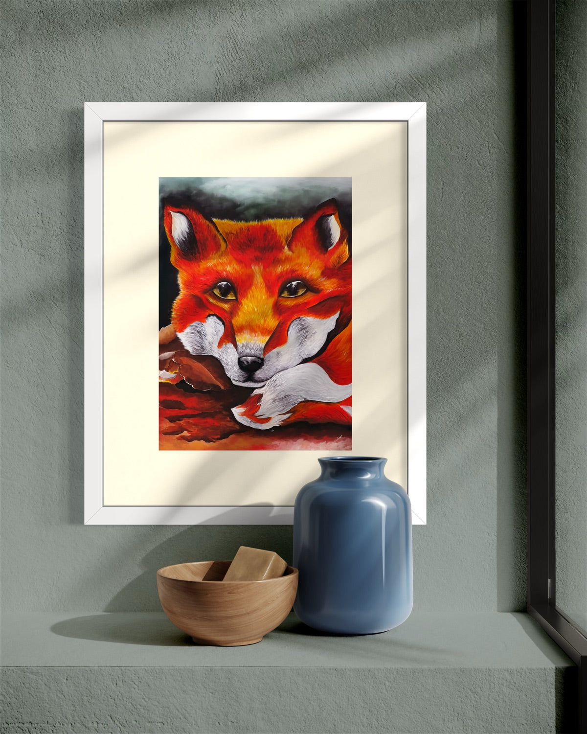 White framed A4 red fox art print in dappled light on a sage grey plaster wall above a built-in shelf with a wooden bowl and blue vase.