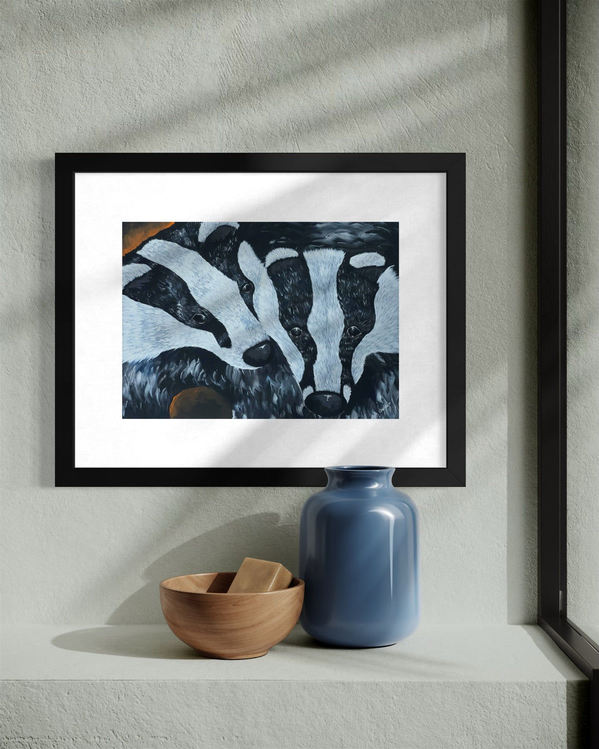 Black framed A4 badger painting in dappled light on a sage plaster wall above a stone shelf with a wooden bowl and a slate blue glass vase.