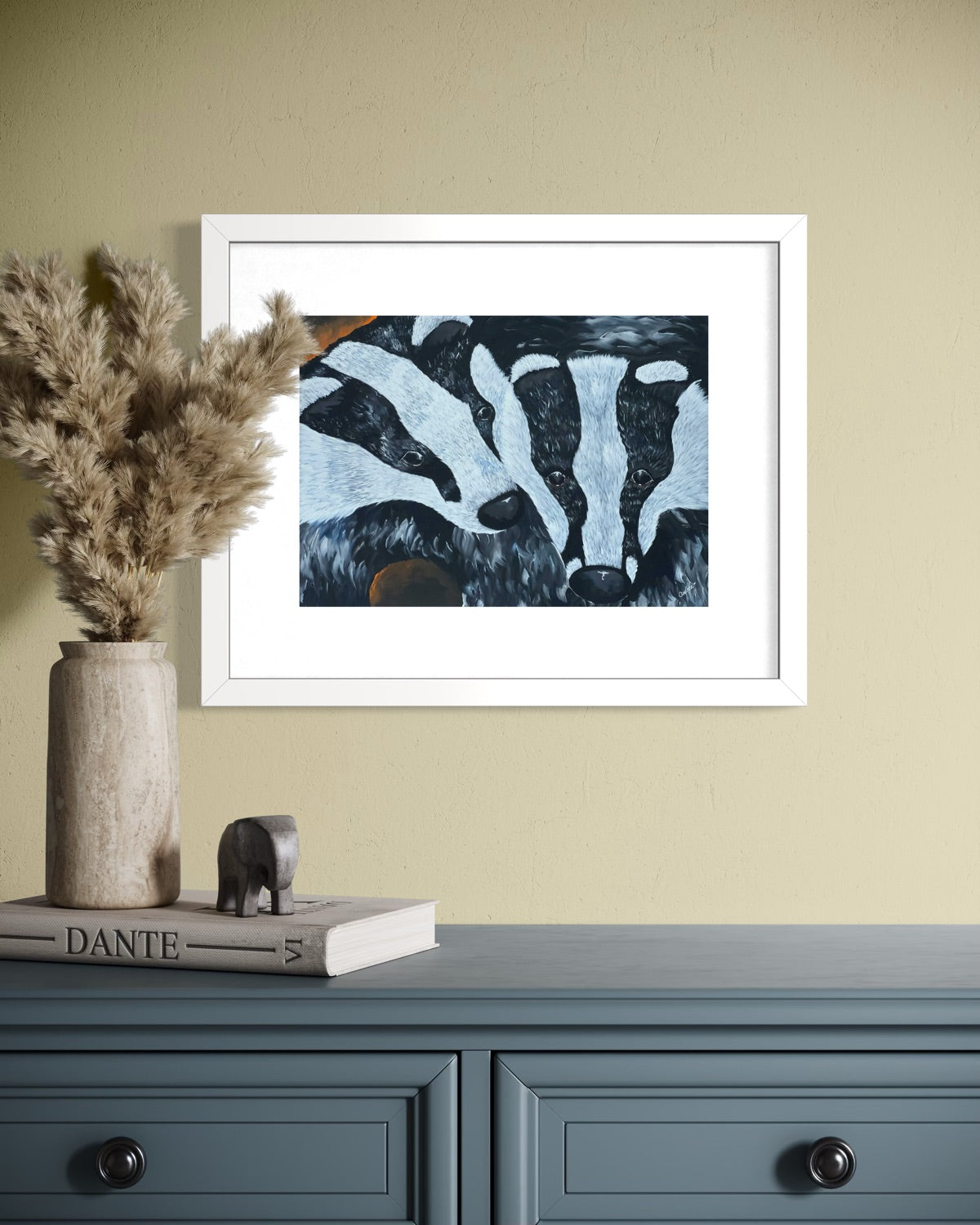 White framed A4 badger art print on a pale yellow plaster wall above a blue grey dresser with pampas grass in a travertine vase and a Dante book.