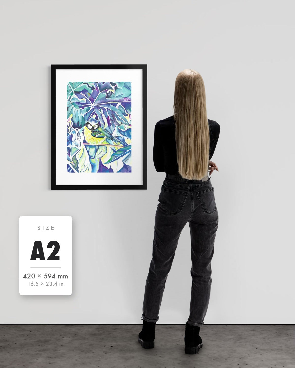 Andrea Nicole 'Taking Flight' blue tit art print in a black A2 frame shown to scale beside a standing woman on a gallery wall, with an A2 (420 × 594 mm) size label. | an:family=print;role=sizeref;size=A2;finish=black