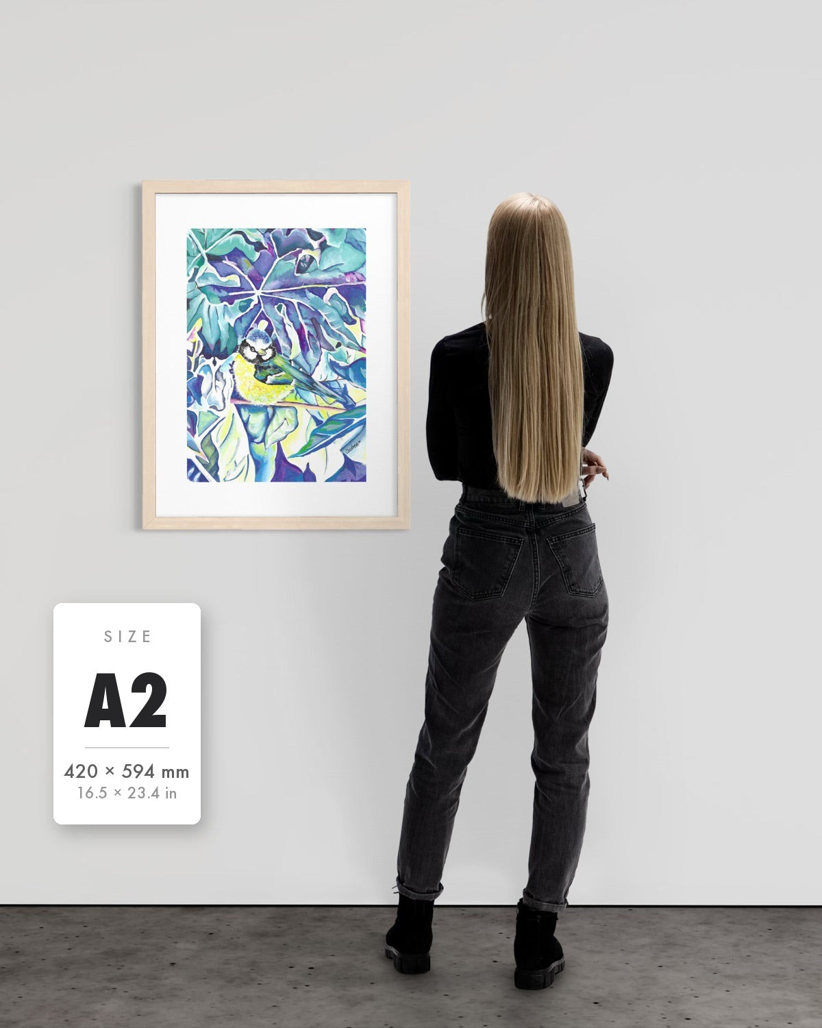 Andrea Nicole 'Taking Flight' blue tit art print in a natural wood A2 frame shown to scale beside a standing woman on a gallery wall, with an A2 (420 × 594 mm) size label. | an:family=print;role=sizeref;size=A2;finish=natural