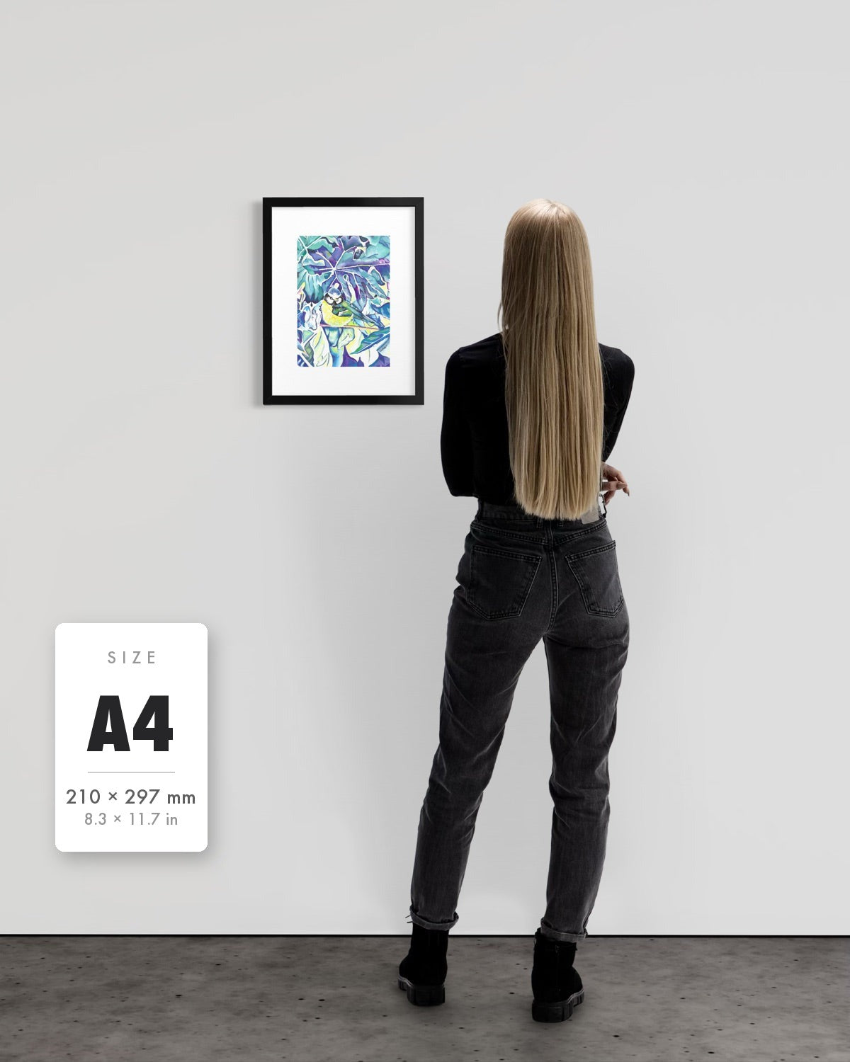 Andrea Nicole 'Taking Flight' blue tit art print in a black A4 frame shown to scale beside a standing woman on a gallery wall, with an A4 (210 × 297 mm) size label. | an:family=print;role=sizeref;size=A4;finish=black