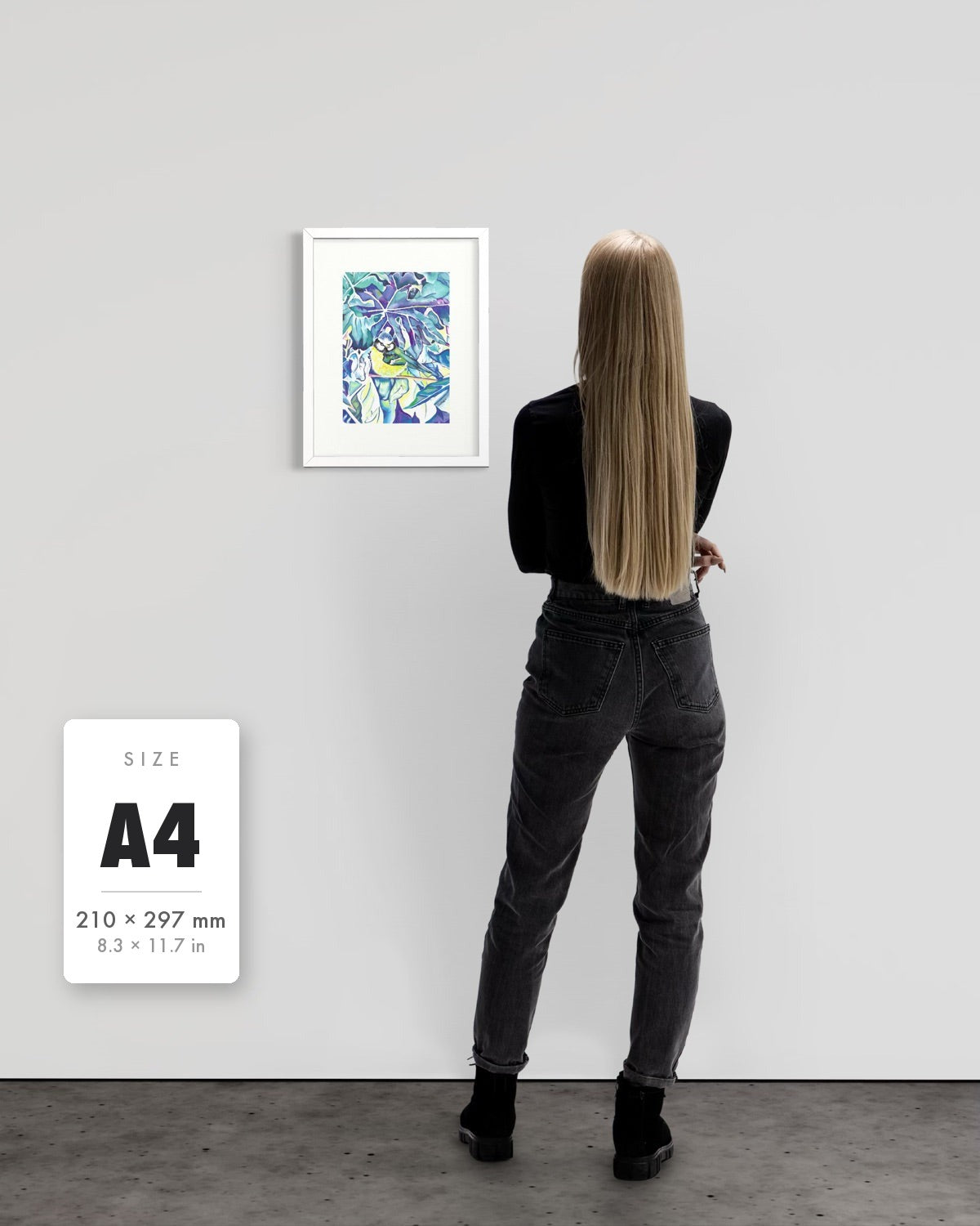 Andrea Nicole 'Taking Flight' blue tit art print in a white A4 frame shown to scale beside a standing woman on a gallery wall, with an A4 (210 × 297 mm) size label. | an:family=print;role=sizeref;size=A4;finish=white