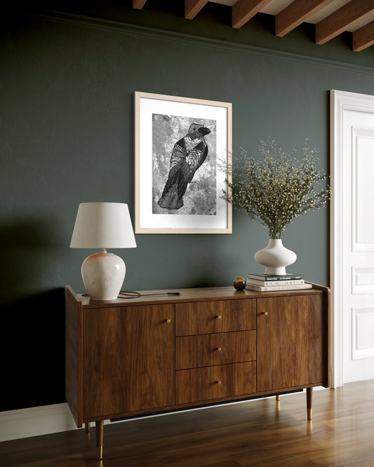 Natural wood framed crow print on a deep teal wall above a walnut mid century sideboard with a white lamp and dried blossoms in a vase.