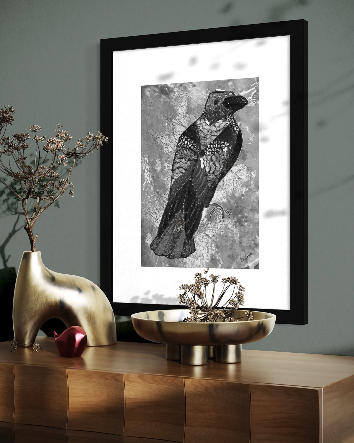 Black framed mixed media crow print on a dark teal wall above a wooden sideboard with brass vases of dried flowers and seed heads.