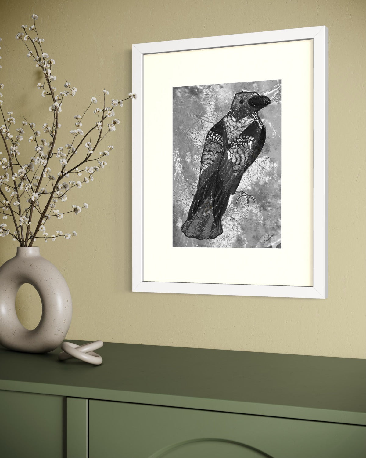 White framed crow print on an olive yellow wall above a green cabinet, with a stone donut vase of pussy willow branches standing beside it.
