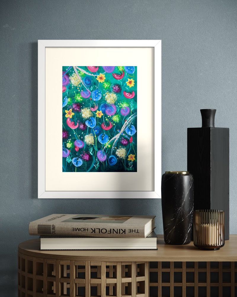 White framed A4 wildflower art print on a slate blue wall above an oak woven side table with the Kinfolk Home book and a marble candle vase.