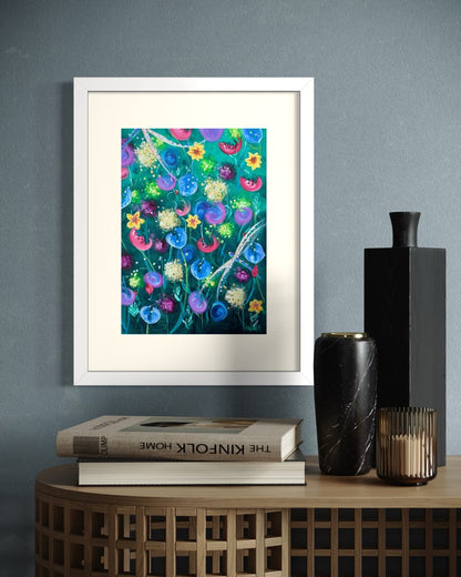 White framed A4 wildflower art print on a slate blue wall above an oak woven side table with the Kinfolk Home book and a marble candle vase.