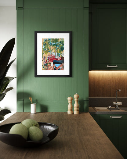Black framed A3 figurative garden art print on sage panelled kitchen walls above an oak counter with a bowl of green melons and pepper mills.