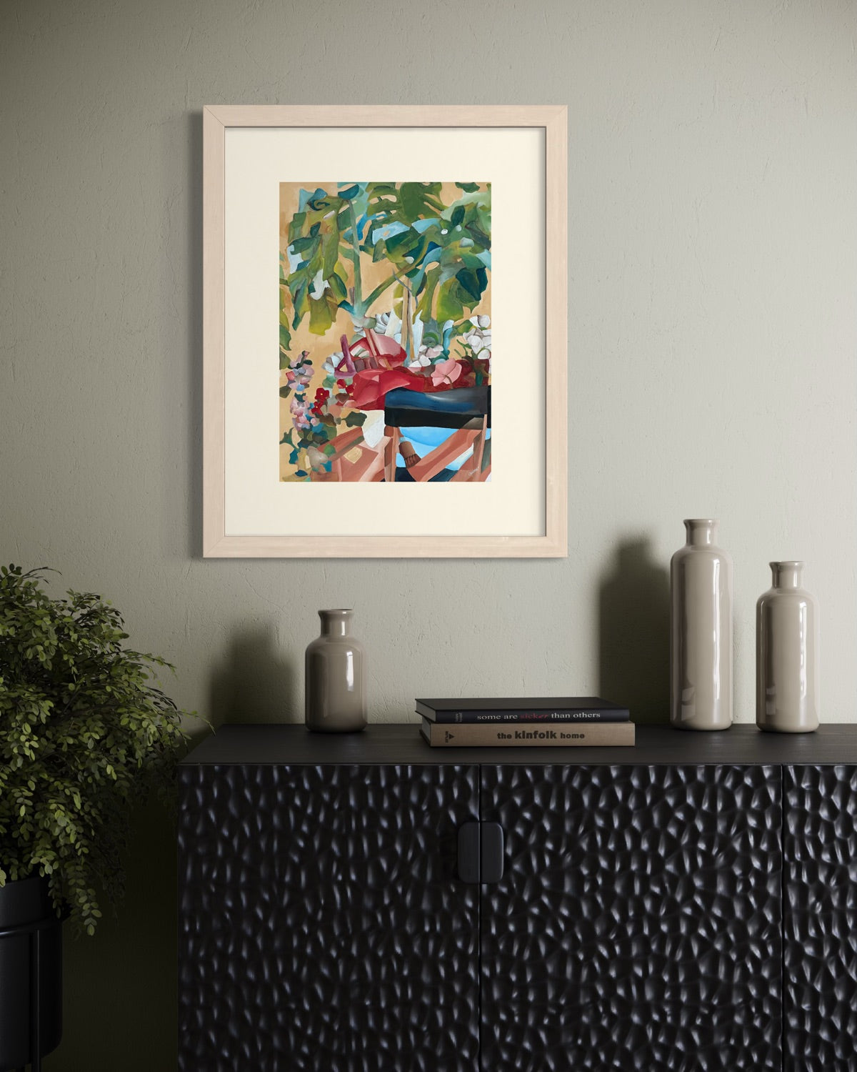 Natural wood A3 garden painting on a sage plaster wall above a hammered black sideboard with sage stoneware bottles and a leafy ficus.