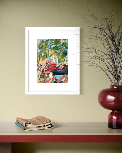White framed A4 figurative garden art print on an olive wall above a red trimmed shelf with leather notebooks and an oxblood gourd vase.